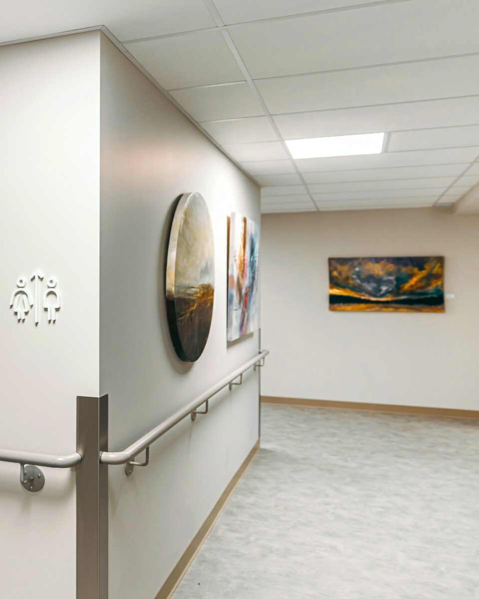 Un corridor et un hall lumineux aux tons blancs, intégrant des œuvres d’art moderne, permettent de créer un environnement apaisant et inspirant dans un centre d’hébergement en cancérologie, favorisant le bien-être et le réconfort des patients.