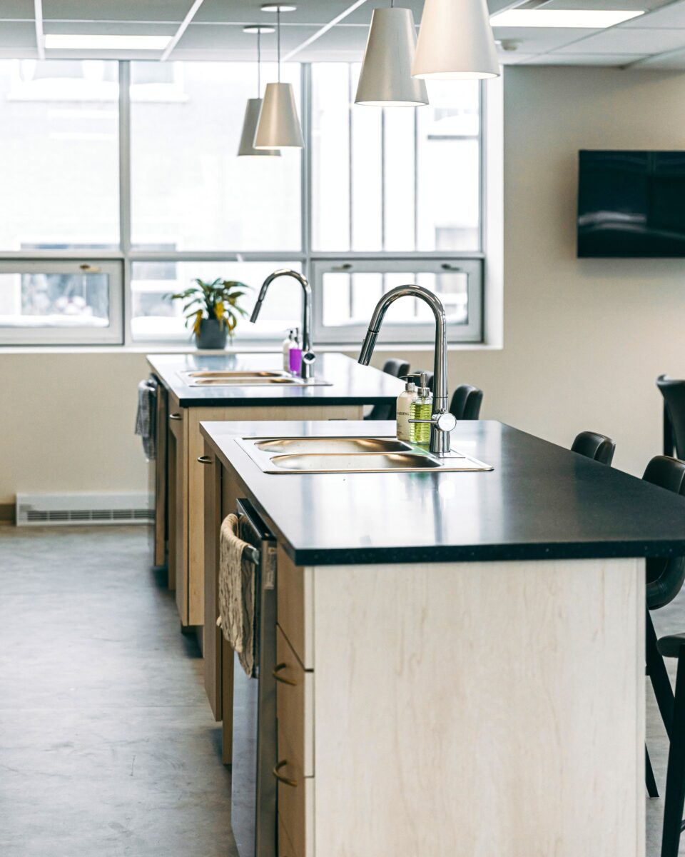 La cuisine et salle commune, lumineuse grâce à de grandes fenêtres, intègre un design moderne mettant en valeur le bois Miralis, un comptoir fonctionnel, un lavabo ergonomique et des luminaires soigneusement sélectionnés, créant un espace chaleureux et convivial.